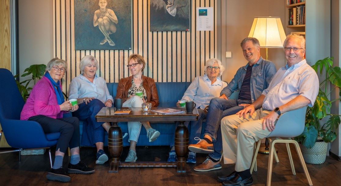 Flere eldre sitter rundt et bord med hver sin kaffekopp i hånda. Noen ser på hverandre, andre ser i kamera.