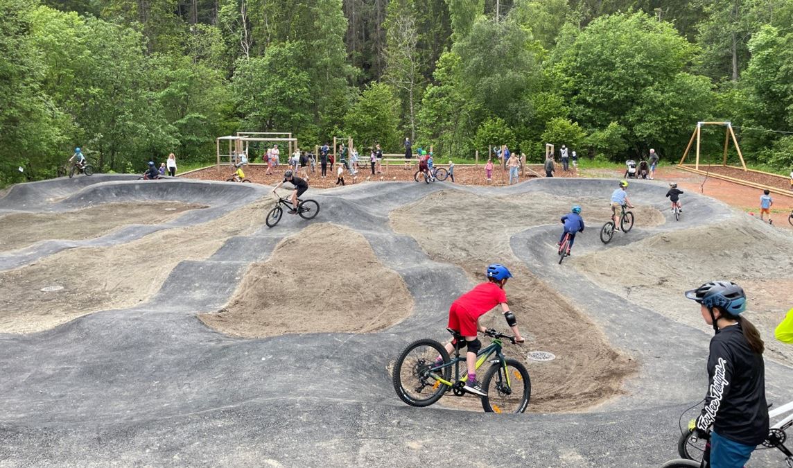 Mange barn på sykler i aktivitet på en sykkelbane. i bakgrunnen ses flere mennesker og en aktivitetspark med ulike treningsaparater.