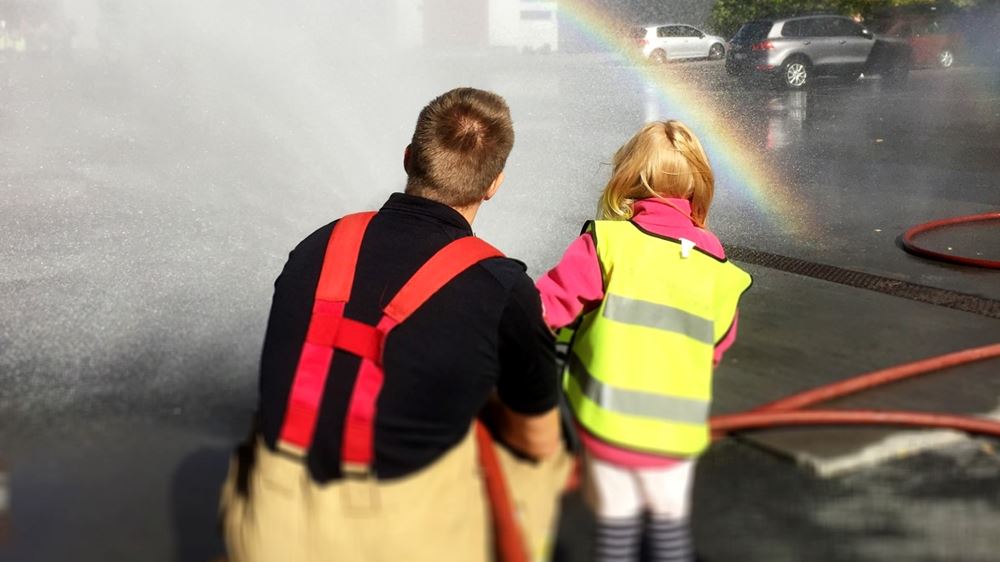 En brannmann sitter på huk med ryggen til kamera, står ved siden av et barn med gul refleksvest. Begge holder i en brannslange som spruter vann.