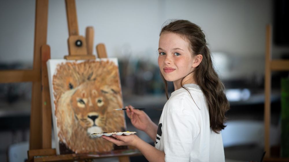 Ei jente står foran et staffeli. Hun er vendt mot kamera, og har pensel og maleskrin i hendene. På staffeliet står et nærbilde av et løvehode.