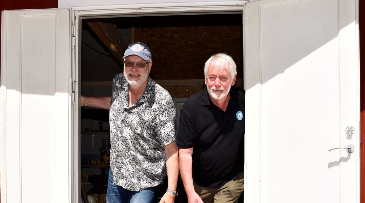 To eldre menn står på dørstokken og titter ut gjennom en åpen dør. Ser mot kamera og smiler.