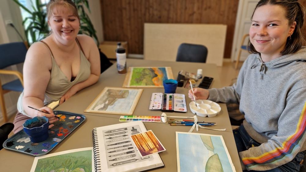 To jenter sitter på hver sin side av et bord, ser mot kamera og smiler. Bordet er fullt med ulike tegninger og malerier, pensler og maleutstyr.