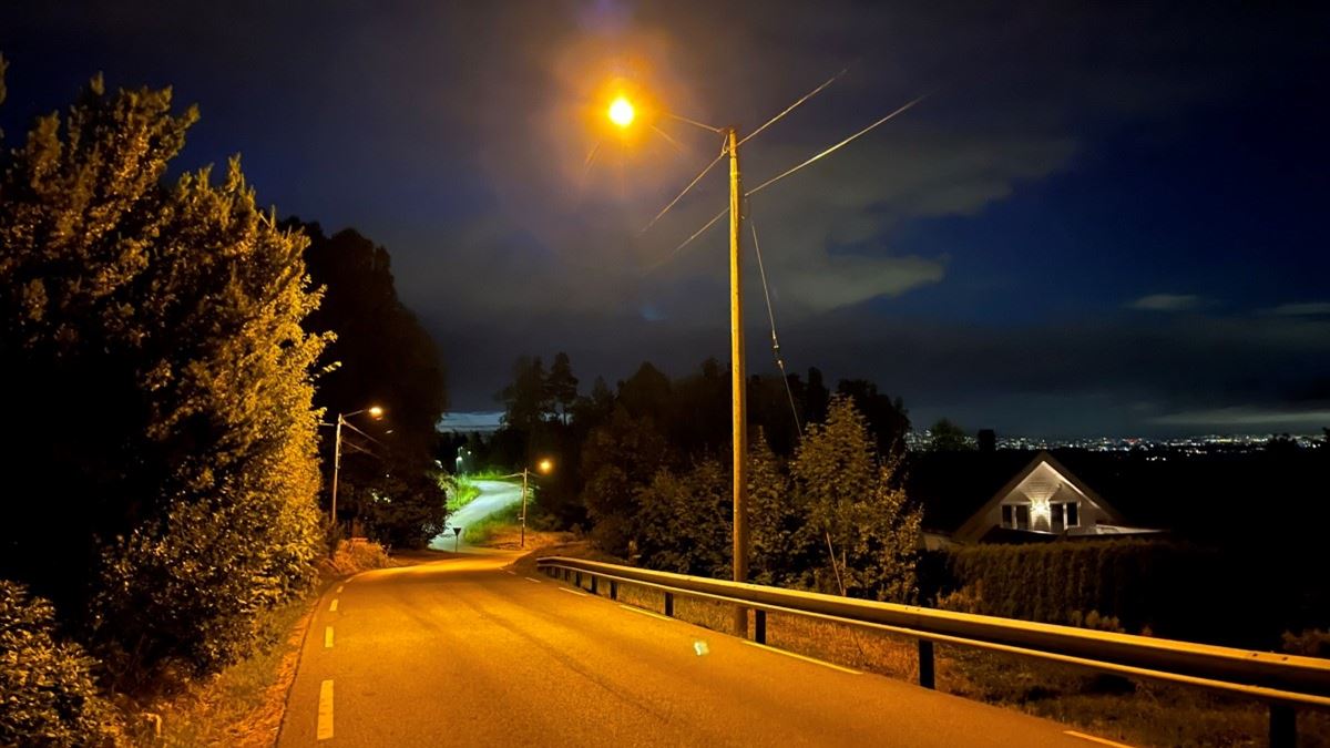 Nattbilde av en opplyst bilvei. Vi ser tre forskjellige lyktestolper som lyser. Det er busker og trær langs veien, og noen hus det lyser fra i bakgrunnen.