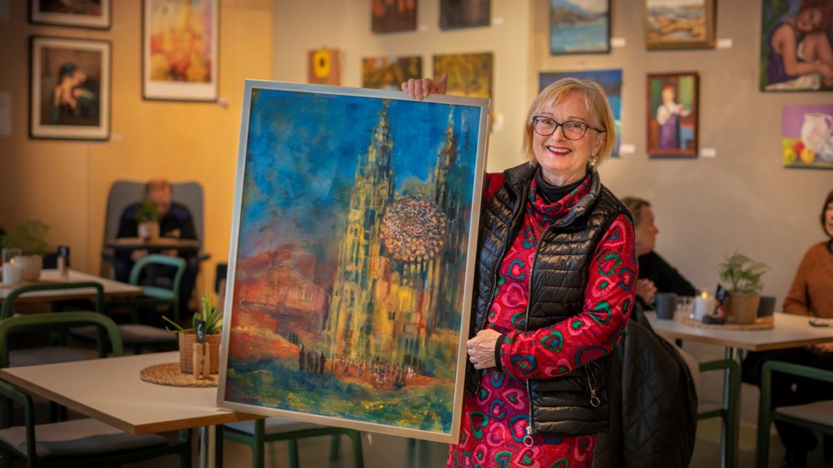En dame står foran en vegg med mange malerier. I hendene holder hun frem et stort og fargerikt maleri, hvor hovedmotivet er en kirke. I bakgrunnen sitter et par personer rundt et bord. 