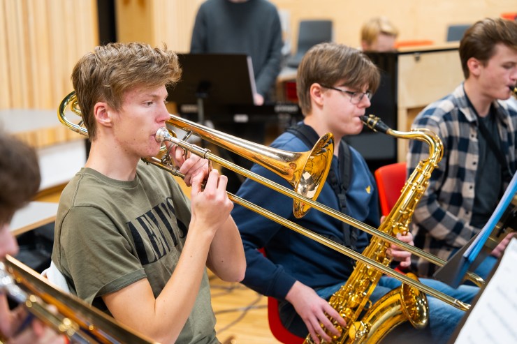 To gutter i fokus sitter ved siden av hverandre i et storband. Den ene spiller trombone, den andre saksofon. I bakgrunnen sitter flere andre i storbandet.