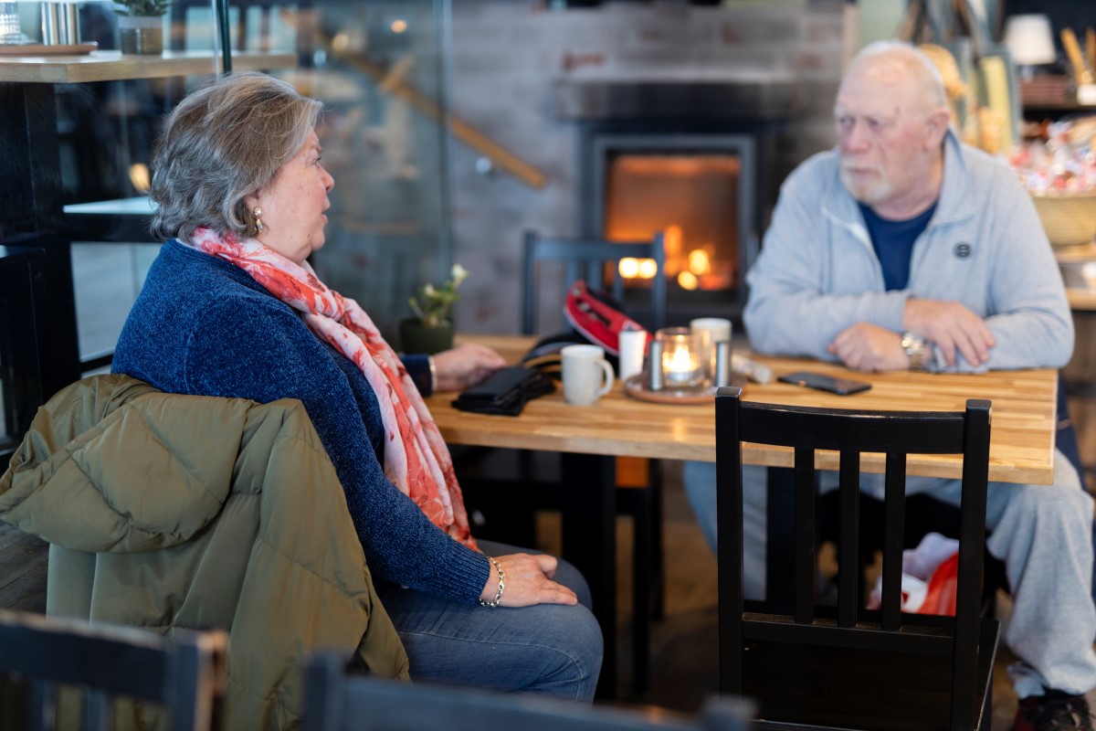 En dame og en mann sitter på hver sin side av et bord, med hver sin kaffekopp. I bakgrunnen brenner det i peisen. 