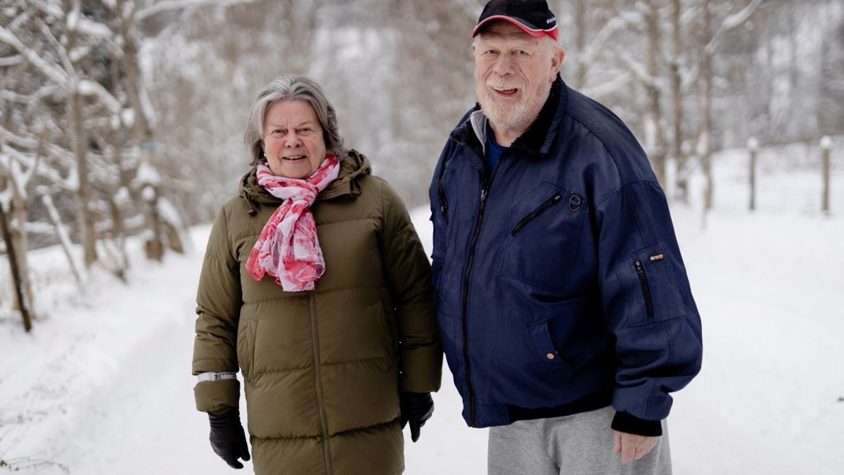 En dame og en mann står ved siden av hverandre på en vei. De er godt kledd med vinterklær, og det er snø i bakgrunnen. Begge ser mot kamera og smiler.