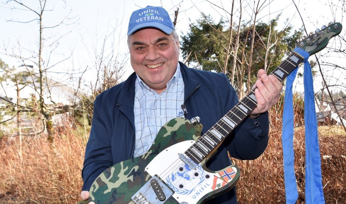 Roy Hovdan smiler og holder opp en gitar med kamuflasjefarger