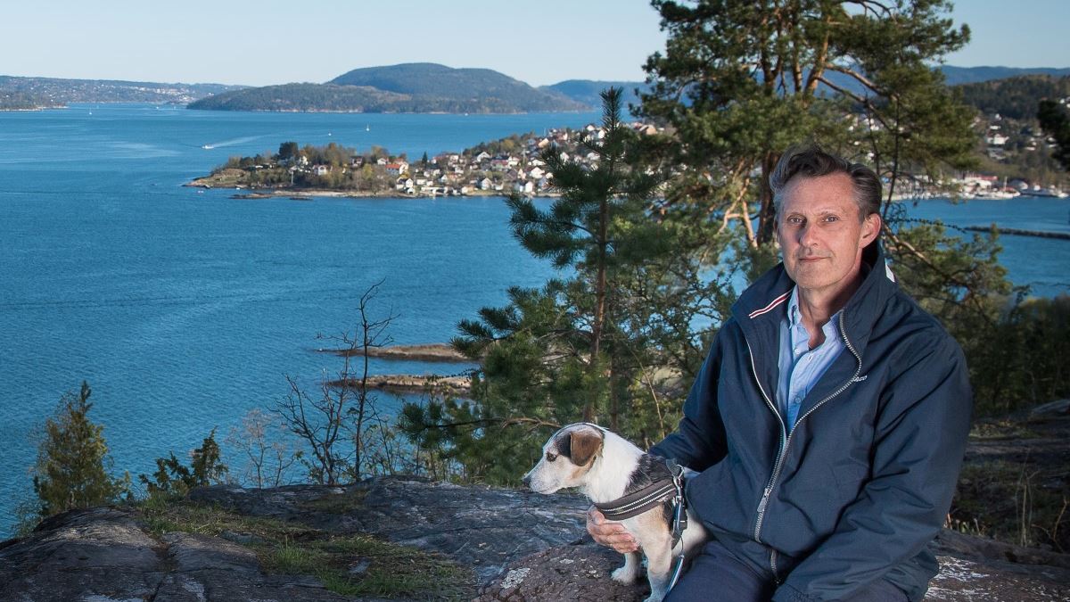 Mann med hund sitter på et utsiktspunkt, trær, sjø og bebyggelse i bakgrunnen