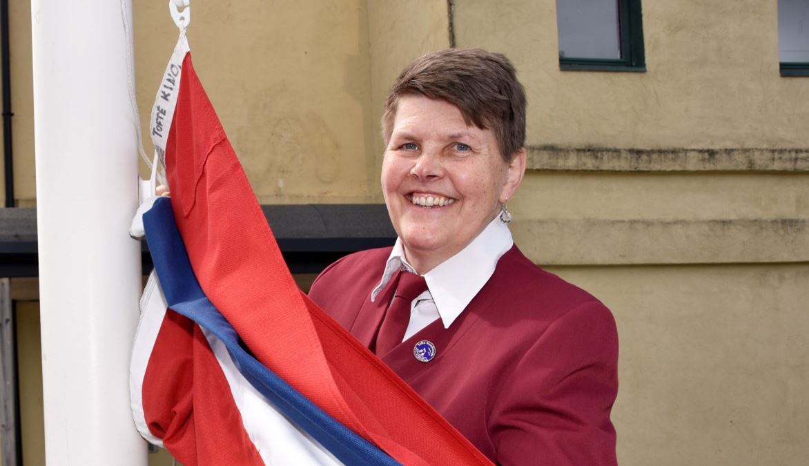 En dame kledd i rød korpsuniform står ved en hvit flaggstang. Holder flagget et norsk flagg i hendene, klar til å heise det.