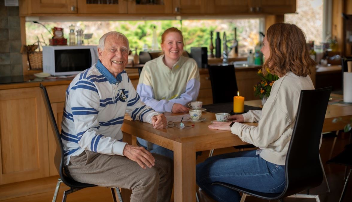 en eldre mann sitter ved kjøkkenbordet sitt, ser i kamera og smiler. Rundt bordet sitter også to yngre kvinner som smiler. I bakgrunnen ses kjøkkenskap, mikroovn og et tent kubbelys.