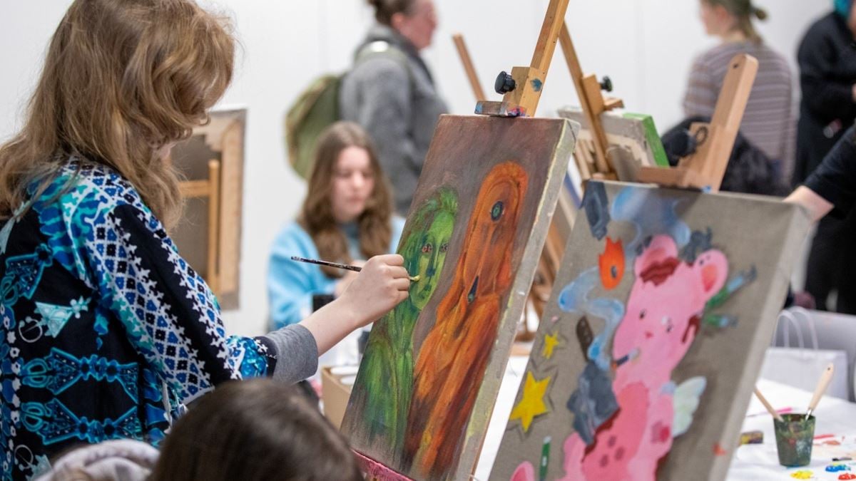 I venstre del av bildet ser vi en jente bakfra. Hun holder en pensel opp mot et lerret, hvor hun maler en grønn person. I bakgrunnen av bildet ser vi flere personer og flere staffelier.