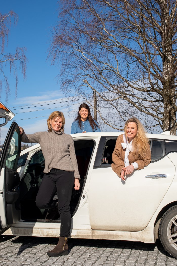 Tre damer titter ut av hver sin dør på en hvit bil. To foran og en ifra baksetet. Døra ved førersetet er åpen, og sjåføren på vei ut av døra.Alle ser mot kamera og smiler.