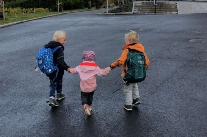 Tre små barn går hånd i hånd, med ryggen mot kamera. De to på hver sin side har skolesekk på ryggen. 
