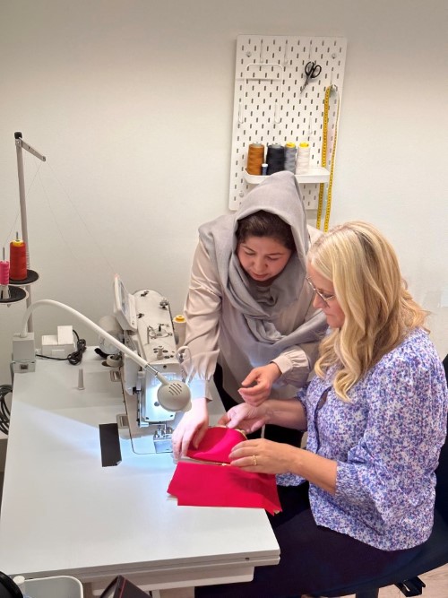 Kronprinsesse Mette Marit sitter foran en symaskin med et rosa stoff i hendene. En annen dame lener seg over fra siden og veileder.