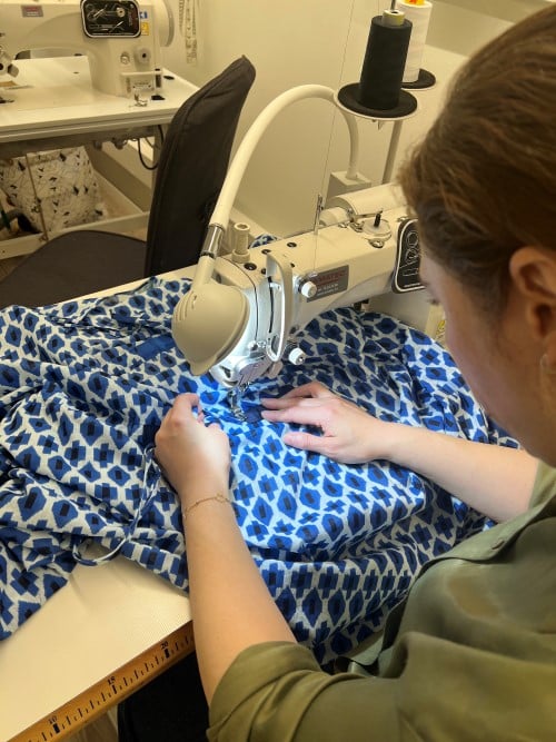 Bildet er tatt bakfra av en dame som sitter ved en symaskin. Under nåla ligger et stort stoff som har mønster i blått, hvitt og svart.
