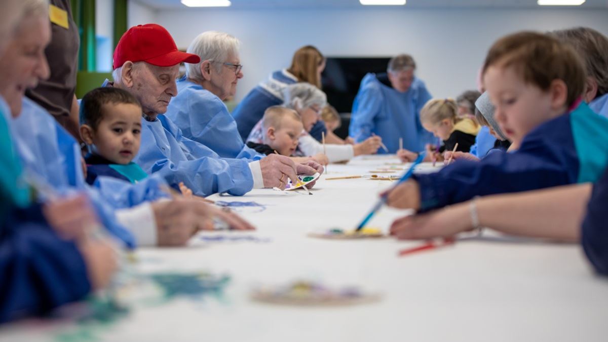 Flere eldre og små barn sitter samlet rundt et langt bord. Alle har blå malefrakker på seg. På bordet er en hvit papirduk. Både små og store har pensler i hånda og maler på duken.