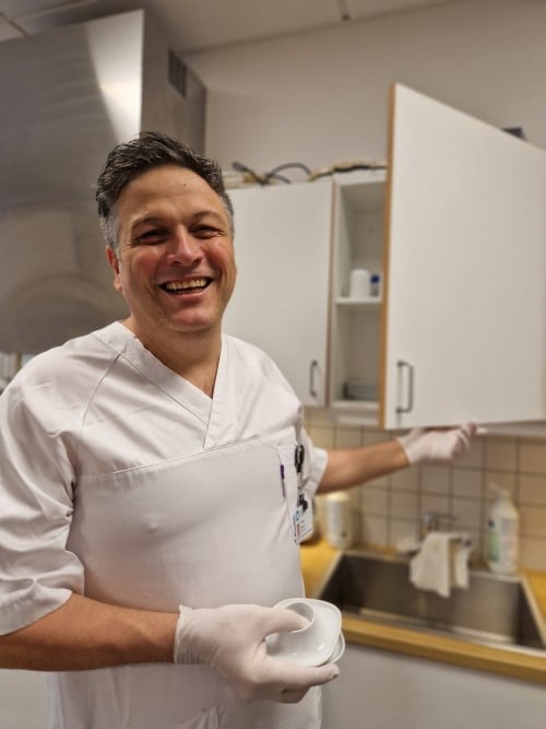En mann i hvit helseuniform setter rene kopper inn i et kjøkkenskap. Han ser mot kamera og smiler.