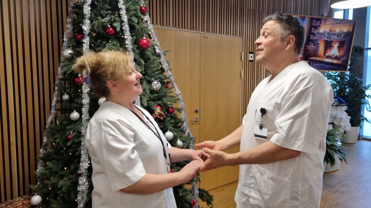 En mann og en dame i hvite helseuniformer holder hverandre i hendene og synger. I bakgrunnen står et juletre.
