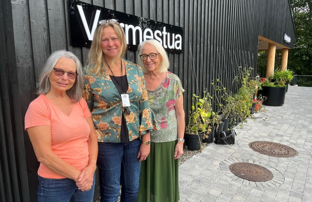 Tre damer står ved siden av hverandre foran et svart hus. PÅ veggen står et skilt med Varmestua.
