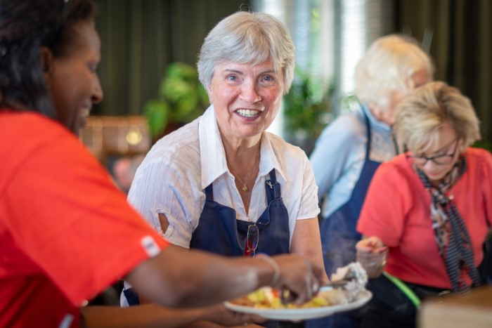 En eldre dame legger opp mat på en tallerken, ser i kamera og smiler.