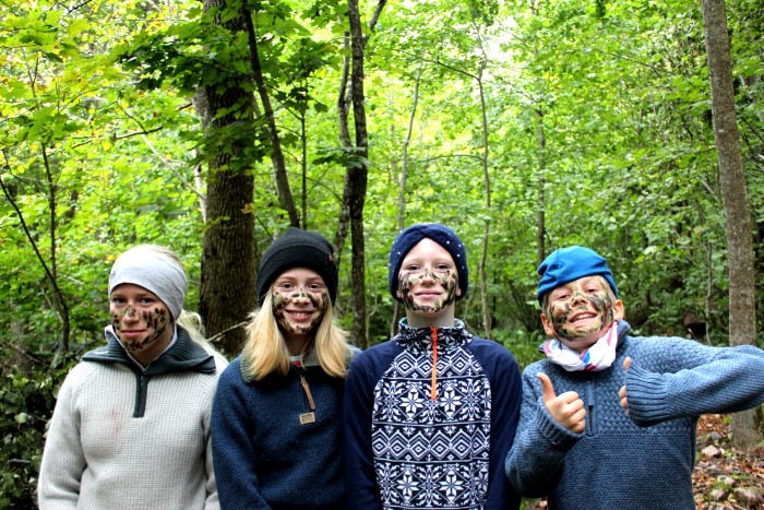 fire barn står ved siden av hverandre i skogen, de har kamuflasjemaling i ansiktet og smiler.