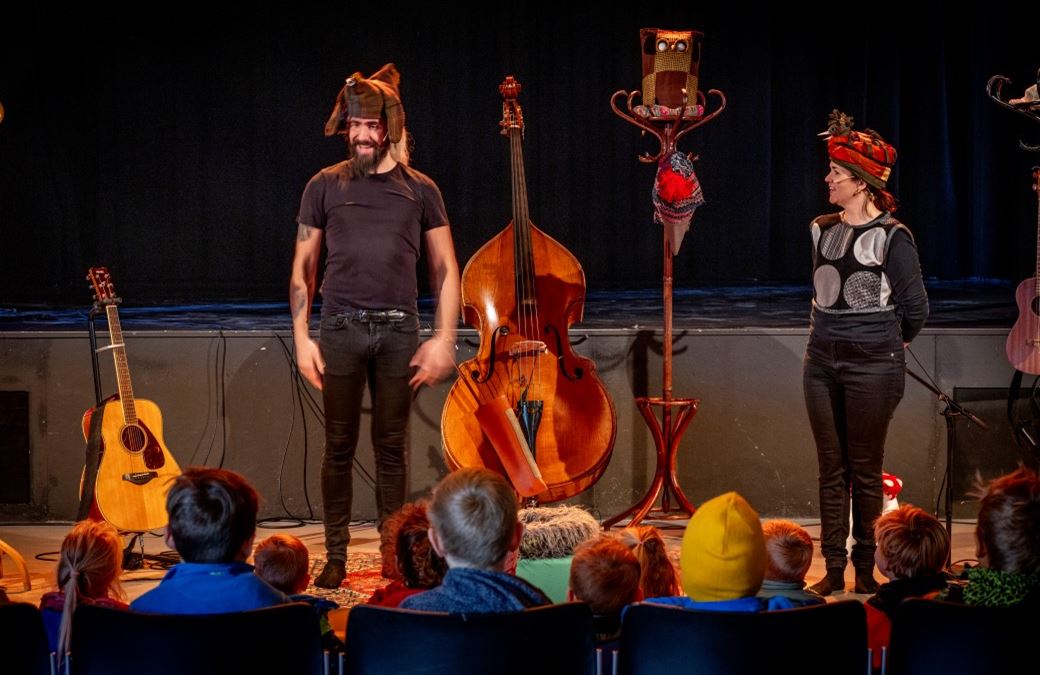 På scenen står to personer med store hodeplagg, en kontrabass og en gitar. I forgrunnen av bildet sitter flere barn, med hodet vendt mot scenen,