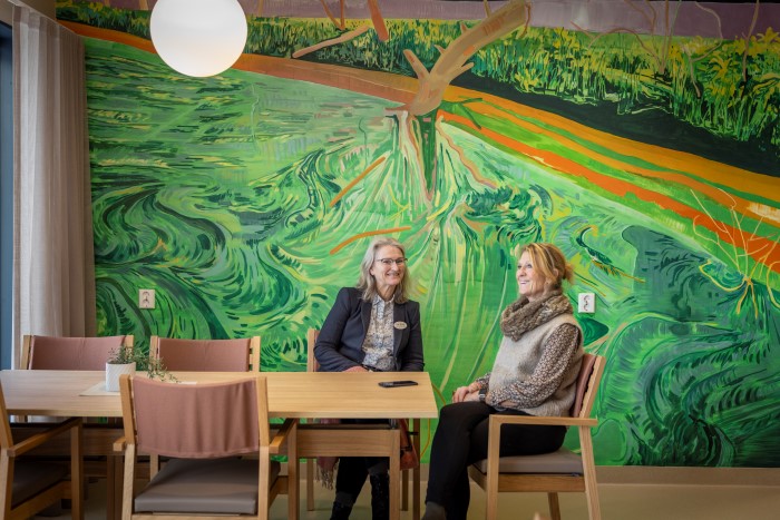 To damer sitter på hver sin side av et bord, ser på hverandre og smiler. Veggen bak dem er dekket av et stort maleri med mye grønt.
