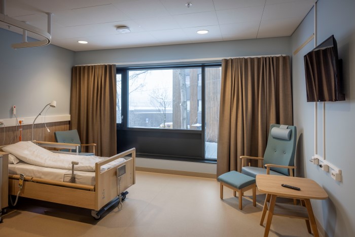 Bilde av et sykehusrom med seng, bord, lenestol, TV og et stort vindu med utsikt.