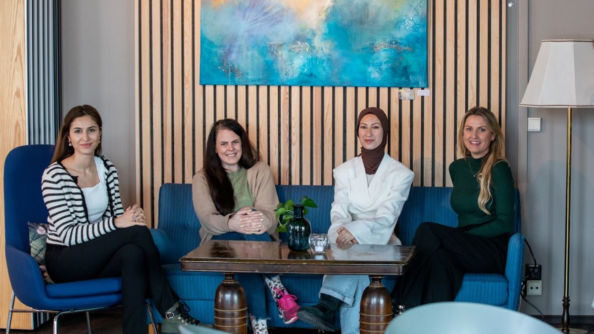 Fire damer sitter i en sofa bak et lavt bord. De ser mot kamera og smiler.