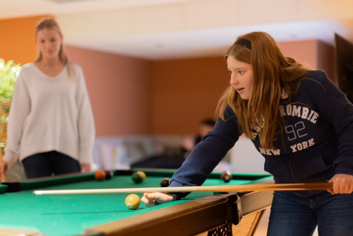 En jente står lent over et billiardbord med en billiardkølle i hendene. På bordet ligger flere kuler. I bakgrunnen står en annen jente.