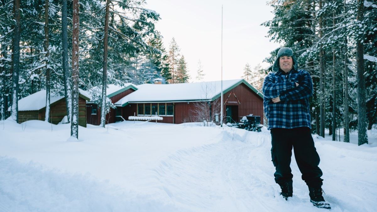 En mann i vinterklær står i forgrunnen av bildet. Det er mye snø, både på bakken og trærne rundt. I bakgrunnen ligger en stor rød hytte.
