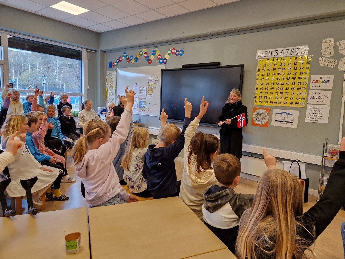 Mange elever i et klasserom rekker hendene i været. Forran ved tavla står en dame med et norsk flagg i hånda og smiler.