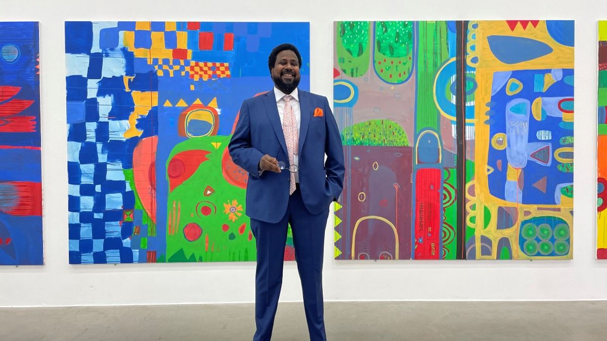 En mann i dress og lakksko står foran en vegg fylt av fargerike malerier i sterke farger. Han smiler.