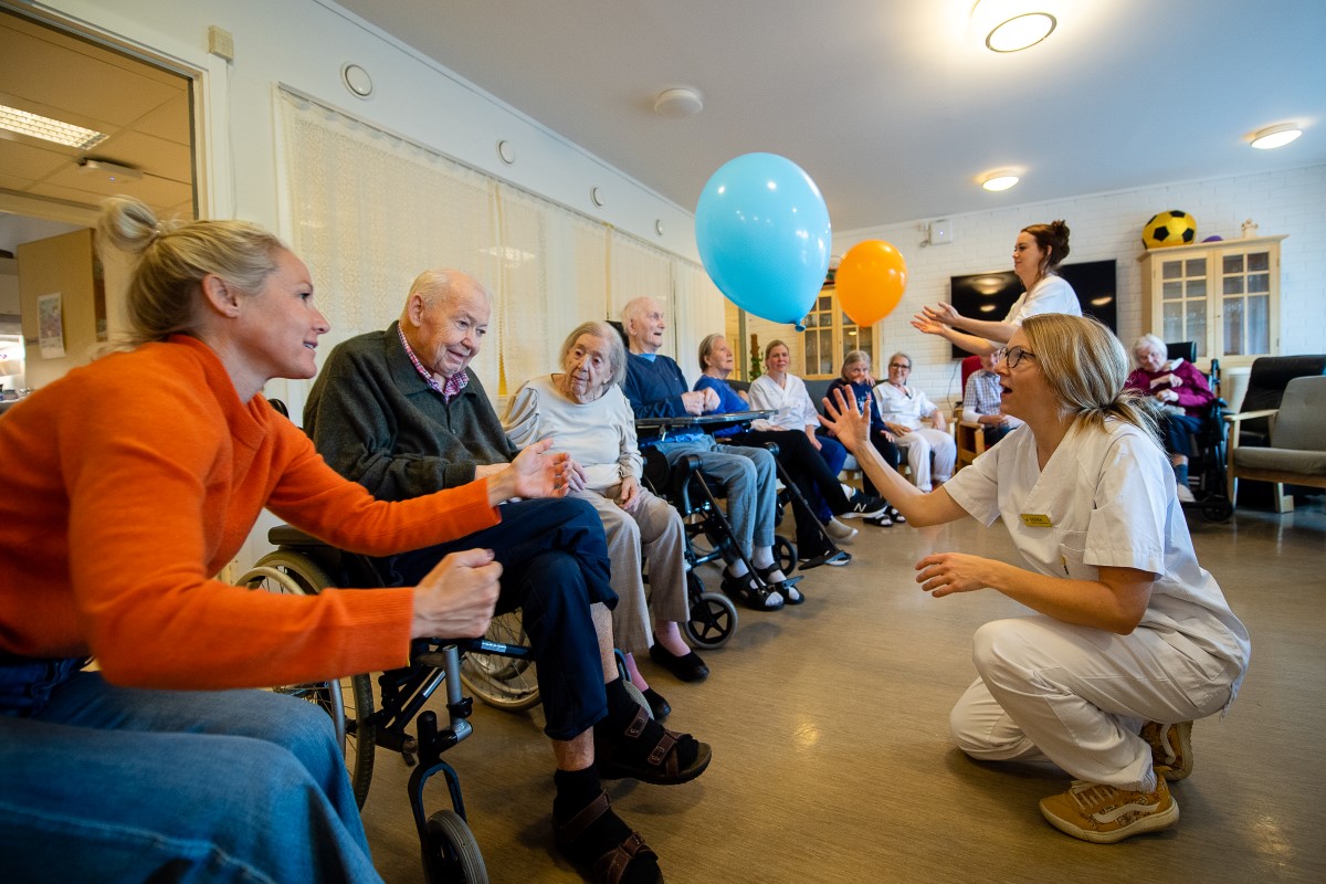 Flere eldre mennesker sitter ved siden av hverandre på en rekke. Noen i rullestol, andre i vanlig stol. De ser foran seg, der to damer kledd i hvitt kaster hver sin ballong opp i lufta.