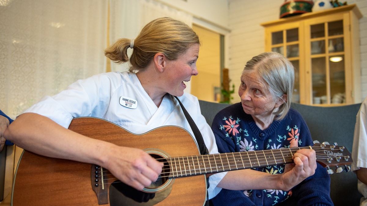 To damer sitter sammen i en sofa. Den ene er kledd i hvite helsearbeiderklær, har navneskilt på brystet og spiller på en gitar. Ved siden av henne sitter en eldre dame. De ser på hverandre, og damen i hvit synger.