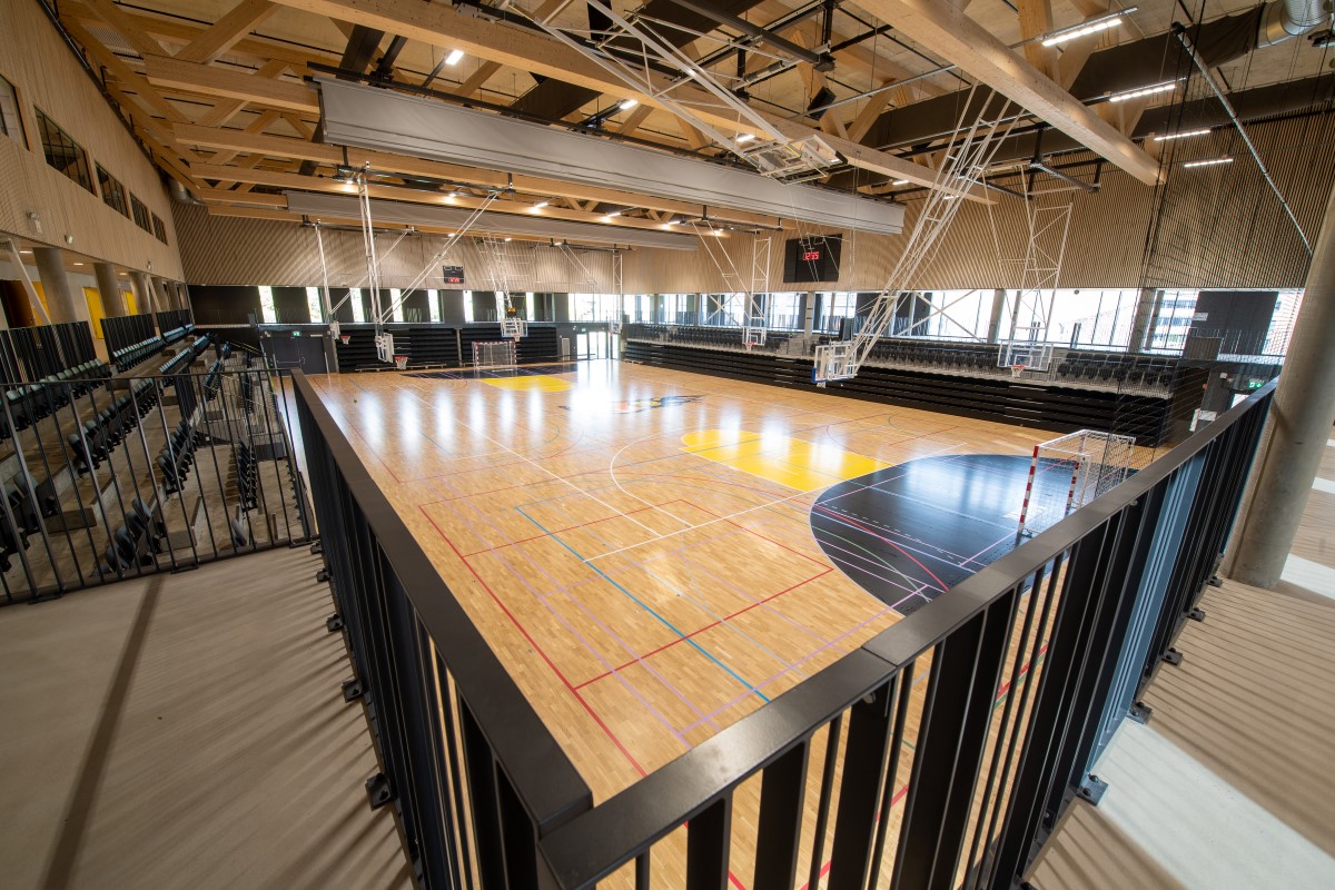 Fra tribunen ser man ned på en innendørs idrettshall med parkettgulv og håndballmål. Langs begge langsidene i hallen er det flere rader med svarte seter for publikum.